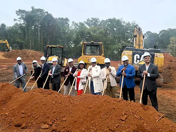 A group of people stand on a construction site holding shovels in the dirt with construction equipment behind them