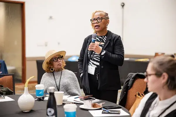 A woman stands and speaks into a hand-held microphone with two people seated on either side of her  