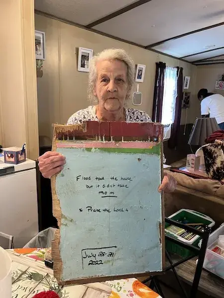 Elder woman holding sign that says Flood Didn't Take Mom