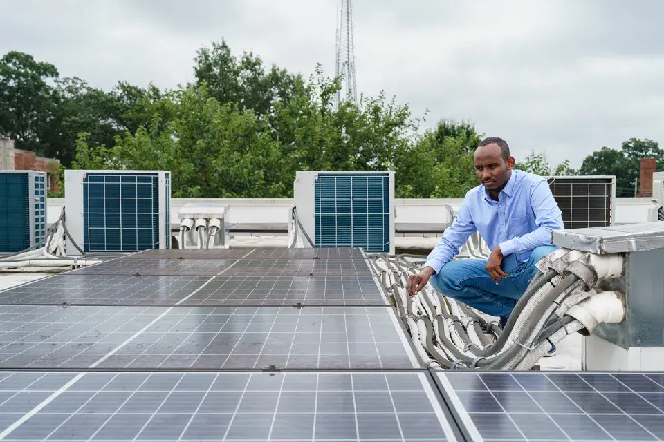 Person in blue dress shirt and jeans with short black hair kneels in front of a solar installation