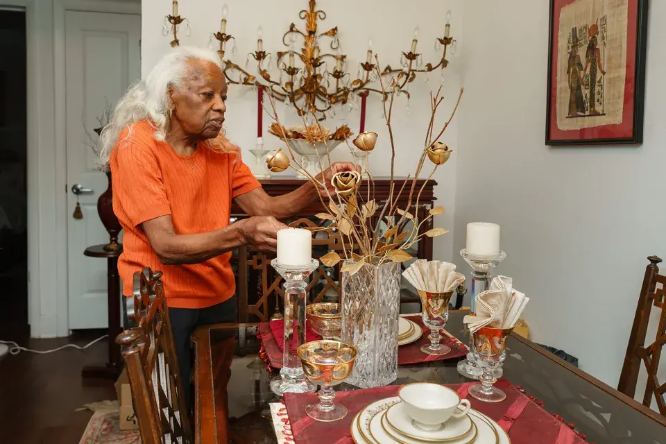 Person with long silver hair and orange top stands in dining room adjusting flower arrangement