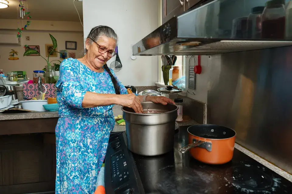 Person with long gray hair pulled into a braid, wearing a blue dress, stands at stove stirring a large pot.