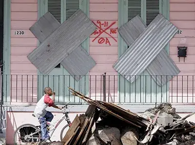Boy on a bicycle riding past a home with boarded up windows and debris piled up on the sidewalk
