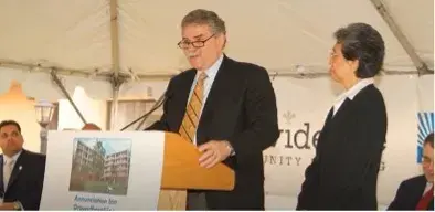 A man at speaks at a podium as a woman stands next to him with other people seated behind them