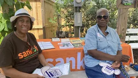 Two women sitting on chairs in front of a table with an orange tablecloth and papers on it
