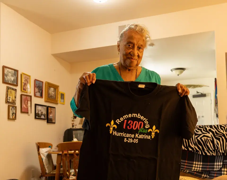 Emelda Paul holds a black t-shirt with the words Remembering Hurricane Katrina bordered by two crosses in a room with framed pictures on the wall
