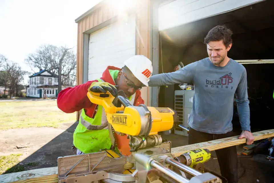 Young person in hardhat uses circular saw while an adult in a blue long-sleeve shirt supervises