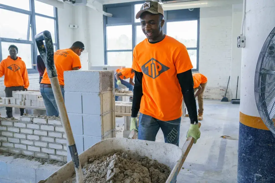 Young person wearing an orange shirt with the logo "TIP" on it wheels a barrel of mortar inside a workshop setting, with other people in orange shirts in the backgrond laying bricks