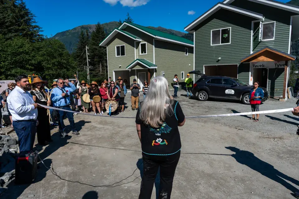 Person with long silver hair wearing an embroidered vest stands in front of a ribbon facing new singl-family homes with trees, mountains, and a blue sky in the background