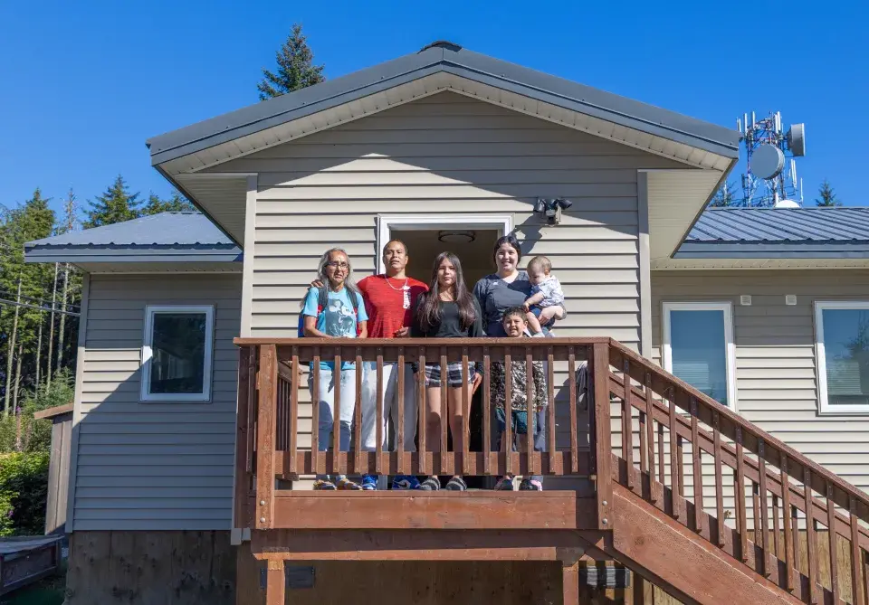 Family stands on the porch of their home with a blue sky and tall trees in the background