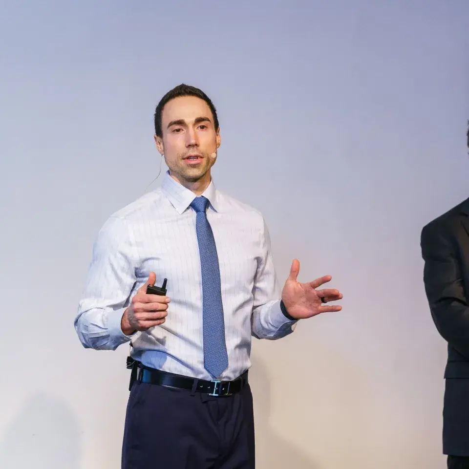 Person wearing light blue shirt and darker blue tie and headset mic speaks gesturing with his hands