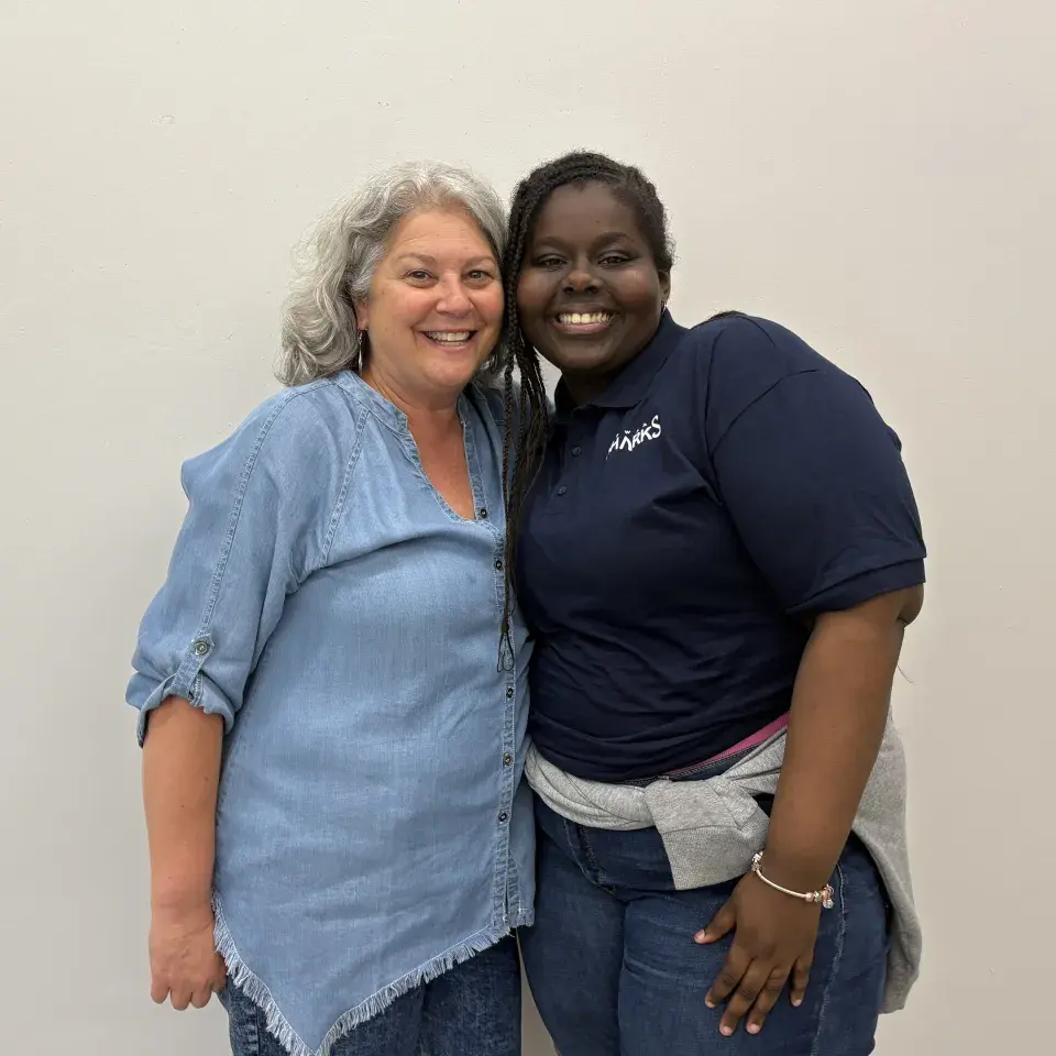Two people stand close to each other smiling; one wears a light blue shirt; the other a dark blue shirt