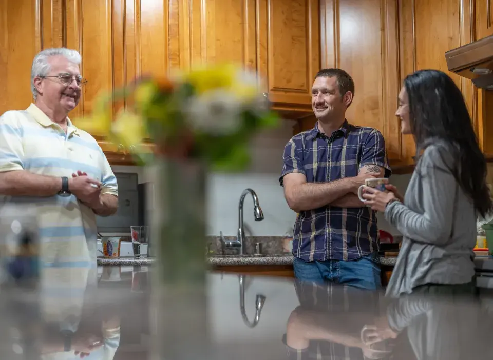 Three people standing in a kitchen at a sink with wooden upper cabinets and a kitchen island with flowers blurred in the foreground