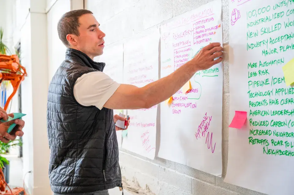 David Goldstein of Hydroshell, 2023 HABC winner, puts a Post-It-Note on one of the whiteboard papers with writing on it lined on a wall