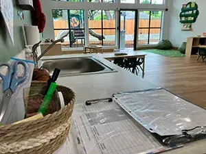 A clipboard, papers, and a basket of supplies rest on a countertop in the foreground of an open classroom with a play area in the back by a wall of windows and door that opens to an outdoor area