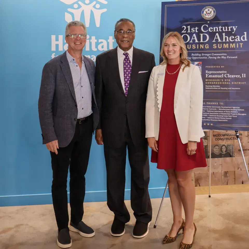 Three people in business attire stand in front of Habitat for Humanity logo and a sign reading, "21st Century ROAD Ahead"