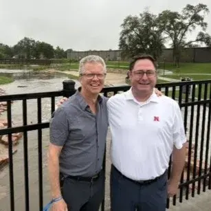 Two people stand smiling with their arms round each other's shoulders and a fence and body of water in the background