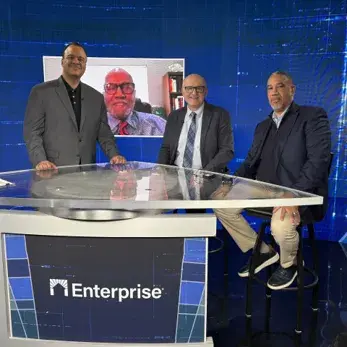 Three people at a table in a newsroom-like setting with another person featured on the screen in the background.