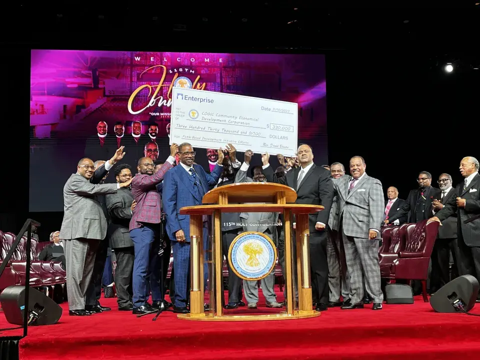 Group of people in business attire stand at a large podium on a red carpet with several holding up a large image of a check.