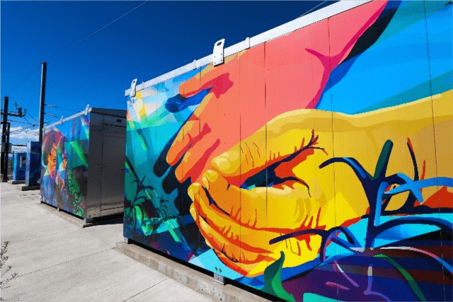 A mural of two different colored hands coming together in the grass and sun. A second mural of people in the background.