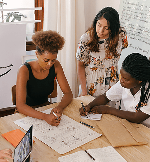 Two women sitting at a table, writing, and discussing a project with another woman standing