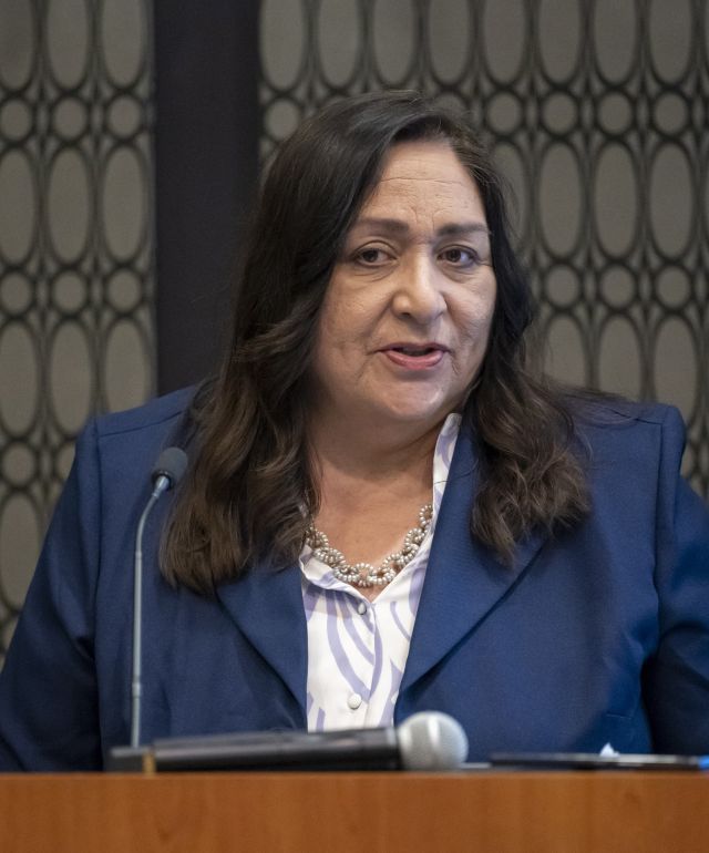 Women with long dark hair wearing a blue suit jacket and white short speaks at a podium.