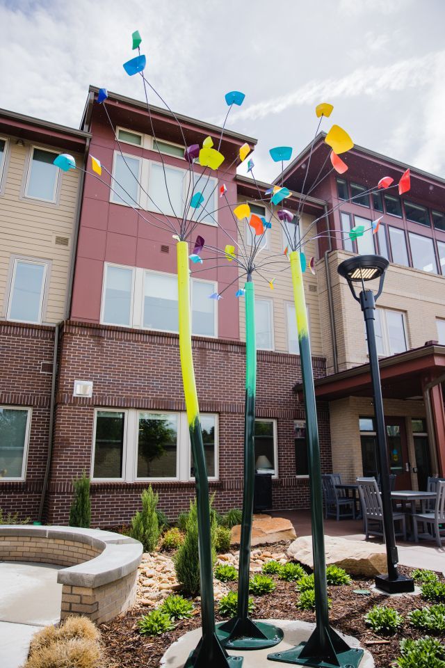 Brightly colored abstract sculpture stands in front of a three story building, next to a lamppost.