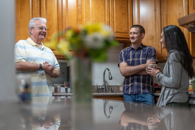 A man with silver hair stands on the left in a kitchen next to a man with short hair and a moustache and a woman with long black hair wearing a gray sweater and holding a mug. The sink is in the background and a vase of flowers sits on the counter in the foreground.