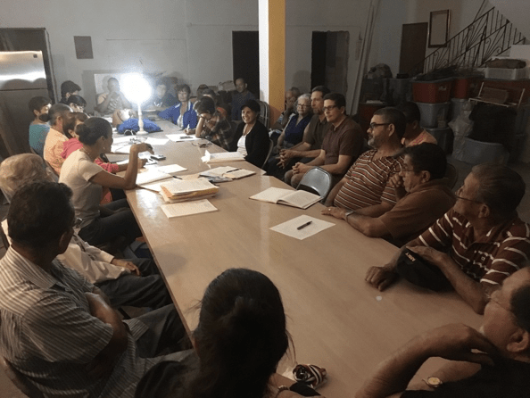 Group of people sit around a long, narrow conference table with papers and files, along with a single lamp providing light.