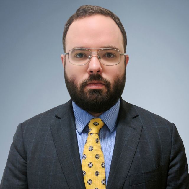 Person with wire glasses and a dark moustache and beard wearing a light blue shirt, a yellow patterned tie, and a dark suit jacket.