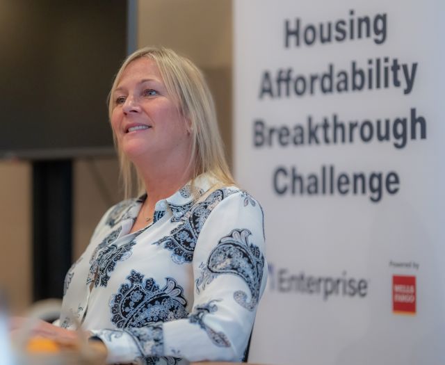 Person with short blond hair stands smiling with signage in the background that says, "Housing Affordability Breakthrough Challenge."