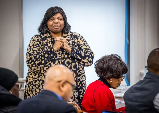 Person with shoulder-length black hair wearing a black and gold patterned dress stands in front of an audience.