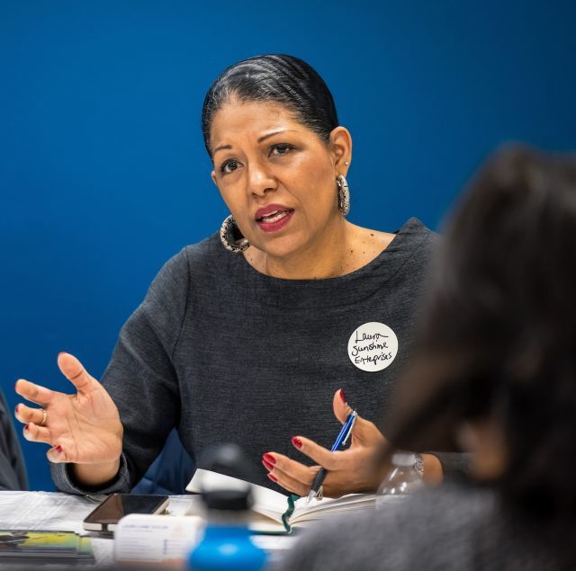 Person with black hair in a low bun wearing silver hoop earrings and a grey scoop neck top is seated at a table, speaking to an audience.