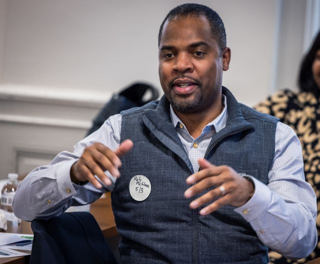 Person with light blue collared shirt and gray fleece vest speaks, gesturing with his hands.