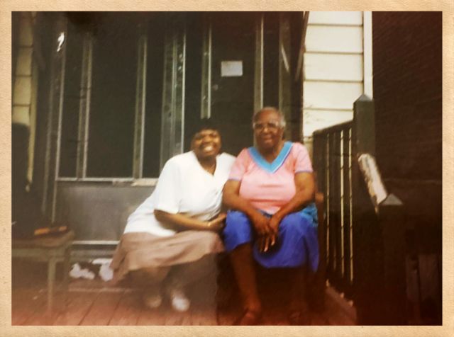 Two people sit on the front porch of a house, side by side, smiling for the camera.