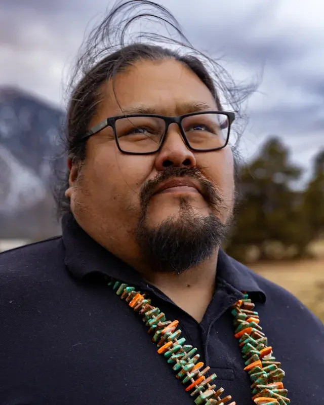Man wearing black square glasses and a beard with beads around his neck looks off into the distance with snow-capped mountains in the background