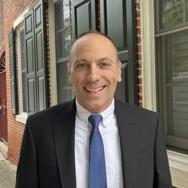 Person smiling in white collar shirt, blue tie and black jacket in front of window panes and brick building