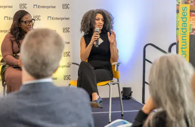 Two people seated on stage with person on right talking and holding a mic