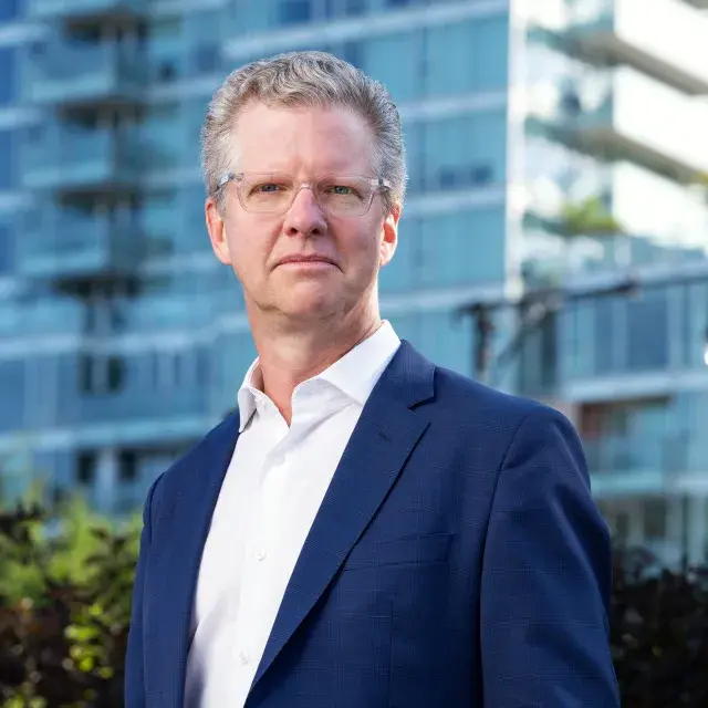 Person with glasses wearing white chirt and blue blazer stands in front of large residential building