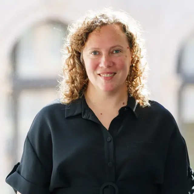 Person smiling with red curly hair and wearing a black blouse; arched windows in the background