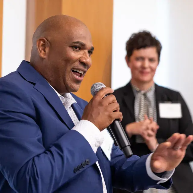 Person smiling and speaking into a mic, gesturing with his hand
