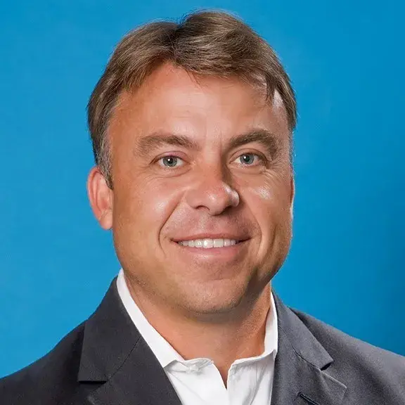 Person smiling with short light brown hair wearing a dark suit jacket and a white collar shirt.