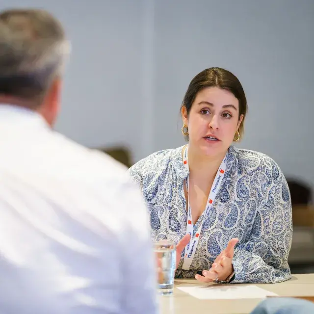 Peson with paisley pattern blouse sitting at a table, speaking and gesturing with their hands