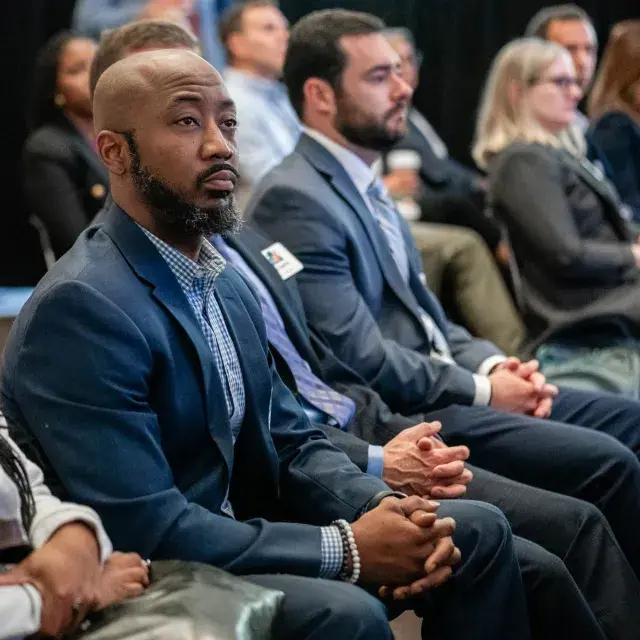 Person with beard wearing blue jacket and collar shirt sits in audience with hands crossed 