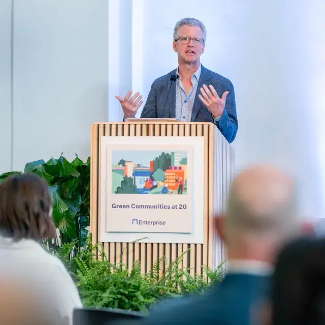 Person with blue shirt and jacket wearing glasses speaks at podium with sign that reads Enterprise Green Communities at 20