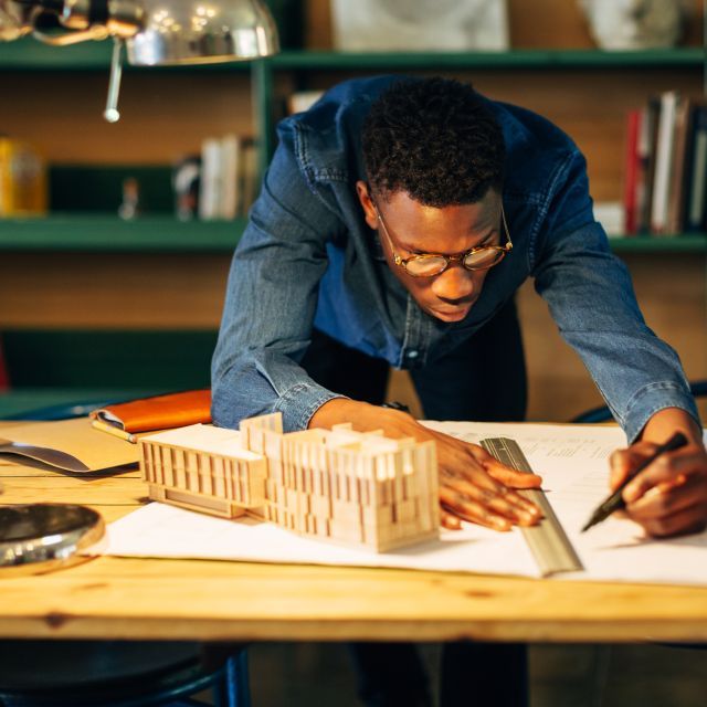 Man leans over wooden drafting table, working on an architectural model and designs