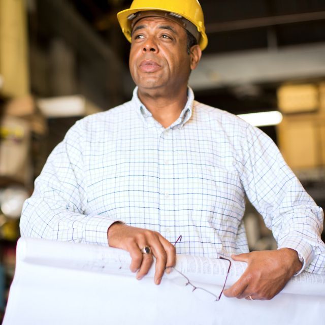 Construction professional in a white dress shirt wearing a yellow hard hat and holding construction drawings.