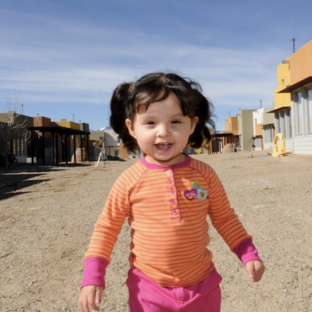A little girl with an orange shirt smiling while standing on a construction site.