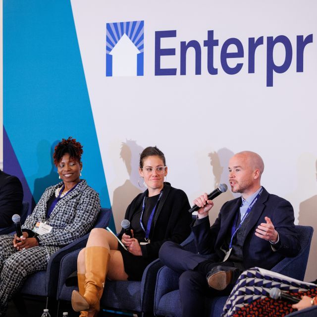 Five people sitting in chairs on stage having a discussion. The Enterprise logo is on the wall behind them. A man holds a microphone.