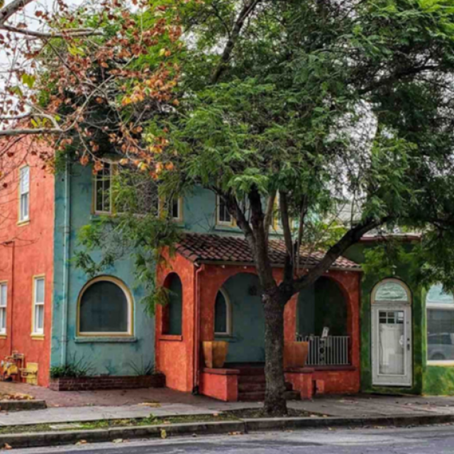 Street view of a small, brightly painted two-story housing property with a tree in front of it. 